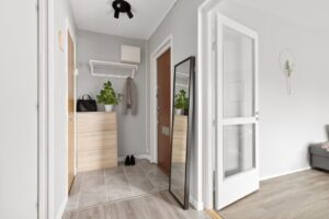 Modern hallway with light wood furniture, a large floor mirror, coat rack, potted plant, and gray tile flooring. An open door leads to a room with wood flooring.