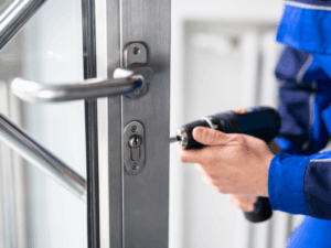 A person in blue work clothes uses a power drill to install or repair a lock on a metal door.