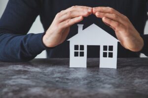 A person’s hands are curved protectively over a small paper cutout of a house resting on a table.