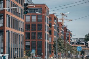Modern red-brick office buildings line a city street with traffic lights, street signs, and construction cranes visible in the background.