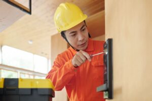 Person in an orange uniform and yellow hard hat uses a screwdriver to install or repair an electronic door lock; a toolbox is visible in the foreground.