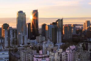 City skyline at sunset with sunlight reflecting off a glass skyscraper, surrounded by other high-rise buildings under a partly cloudy sky.