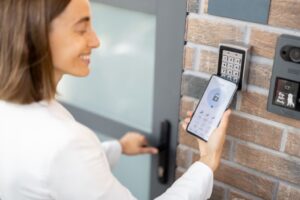 A woman using a smartphone to unlock a door with a keypad access system, while opening the door with her other hand.